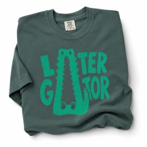 Blue Spruce Comfort Colors folded graphic tee featuring a playful Later Gator alligator illustration forming the letter A in chunky retro green typography.