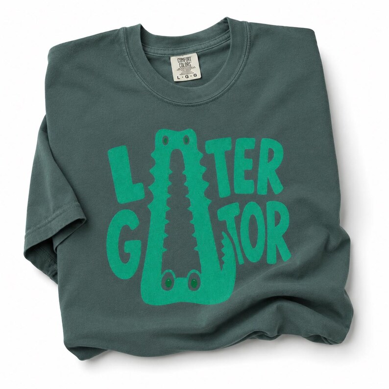 Blue Spruce Comfort Colors folded graphic tee featuring a playful Later Gator alligator illustration forming the letter A in chunky retro green typography.