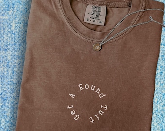 May include: A brown t-shirt with the text "Just a Round  'bout  get" in a circle on the front. The shirt is made of a soft, comfortable material.