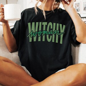 May include: A black short-sleeved t-shirt featuring the text "WITCHY" in large, distressed, light green block letters with the word "woman" written in a cursive, dark green script font layered directly over the center of the design.