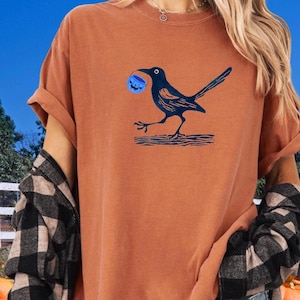 May include: A burnt orange t-shirt featuring a navy blue illustration of a crow carrying a blue pumpkin. The crow is perched on a navy blue log. The shirt has rolled-up sleeves and a relaxed fit.