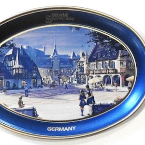 May include: A blue and gold oval metal tray with a picture of a German village scene. The tray has the words "World Showcase" and "GERMANY" printed on it.