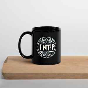Puede incluir: Taza de cerámica negra con un diseño circular blanco con el texto "INTP" en un patrón geométrico.