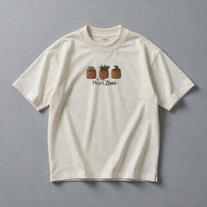 Könnte beinhalten: Ein cremefarbenes Kurzarm-T-Shirt mit einer Stickerei von drei kleinen Sukkulenten in braunen Töpfen. Unter den Pflanzen ist der Text "Plant Lover" in einer dunkelgrünen Schreibschrift gestickt. Das Shirt hat einen gerippten Rundhalsausschnitt.