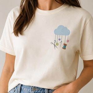 Può includere: Una t-shirt a maniche corte color crema con un ricamo sul petto sinistro. Il disegno raffigura una nuvola azzurra da cui pendono accessori da cucito, tra cui forbici argentate, un ago, un rocchetto di filo e alcuni bottoni.