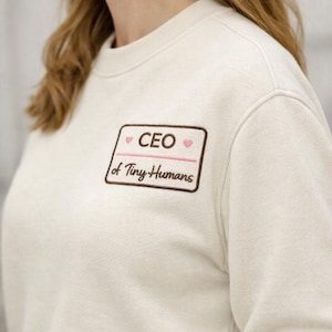 May include: A cream-colored crewneck sweatshirt featuring a rectangular embroidered patch on the chest. The patch has a brown border and contains the text "CEO of Tiny Humans" in brown, accented by two small pink hearts and a horizontal pink line.