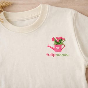 Può includere: Una t-shirt color crema con un ricamo raffigurante un annaffiatoio rosa pieno di tulipani rosa. L'annaffiatoio presenta un piccolo cuore bianco sul lato. Sotto il disegno, il testo "tulipamami" è ricamato in rosa e verde.