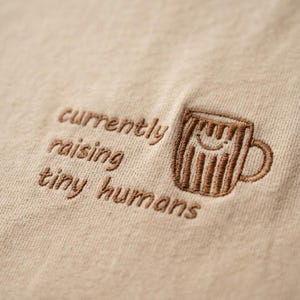 May include: A beige t-shirt featuring brown embroidery. The text reads currently raising tiny humans, accompanied by an embroidered brown coffee mug graphic with a simple smiling face and vertical line details on the surface of the mug.