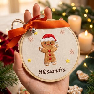 Può includere: Un telaio da ricamo circolare in legno contenente un tessuto color crema con un omino di pan di zenzero ricamato che indossa un cappello da Babbo Natale rosso. Il nome Alessandra è ricamato sotto in marrone. Sono fissati un nastro arancione e un campanellino.