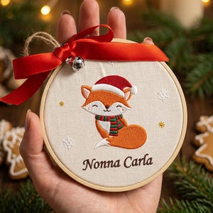 Può includere: Un ornamento rotondo in telaio da ricamo in legno con sfondo in tessuto color crema, ricamato con una volpe arancione che indossa un cappello da Babbo Natale rosso e una sciarpa a righe verdi e rosse. Il testo Nonna Carla è ricamato.