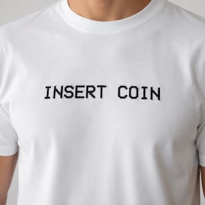Puede incluir: Una camiseta blanca de manga corta con el texto INSERT COIN bordado en hilo negro en el pecho. Las letras están diseñadas con un estilo de fuente pixelada, que recuerda a los gráficos de los juegos arcade clásicos, centradas al frente.
