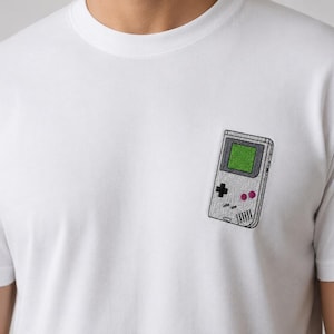 Può includere: Una t-shirt bianca a girocollo con un piccolo ricamo sul petto sinistro. Il ricamo raffigura una console per videogiochi portatile grigio chiaro con uno schermo verde brillante, una croce direzionale nera e due pulsanti circolari magenta.