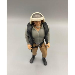 May include: A vintage action figure of a character from a science fiction film. The figure is wearing a white helmet, gray shirt, black vest, tan pants, and black boots. The figure is standing upright with arms at its sides.