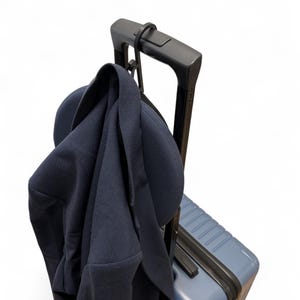 Peut inclure: Une valise bleue avec une poignée noire et un crochet noir attaché à la poignée. Une veste bleu marine est accrochée au crochet.