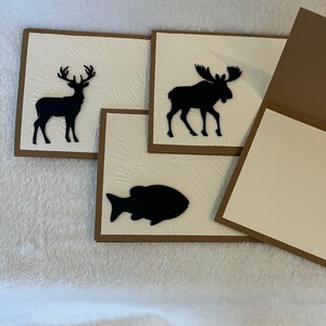 May include: Three brown cards with white interiors. The cards feature black silhouette cutouts of a deer, a moose, and a fish.