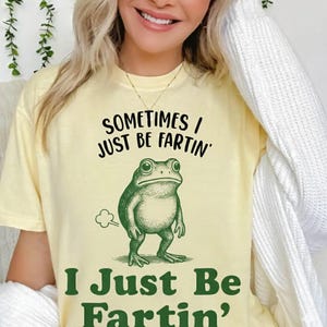 Peut inclure: T-shirt jaune clair avec le texte "SOMETIMES I JUST BE FARTIN'" et "I JUST BE FARTIN'" avec une illustration de grenouille verte. La grenouille est dessinée dans un style vintage, avec un nuage indiquant un pet.