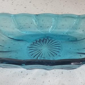 Puede incluir: Un plato de vidrio transparente de color verde azulado con un borde festoneado. El plato de forma ovalada tiene un diseño texturizado de rayos de sol en el centro. El plato mide aproximadamente 25 cm de largo y 13 cm de ancho.