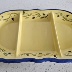 May include: An oval ceramic serving platter with three divided sections. The platter features a pale yellow center with a painted green vine and dark berry pattern, a deep blue scalloped rim, and two light brown scroll-shaped handles on each side.