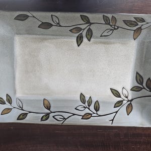 May include: Rectangular ceramic serving platter with a light green border and a central cream-colored area. The platter is decorated with a delicate vine and leaf design in shades of brown and green, adding a touch of nature-inspired elegance.
