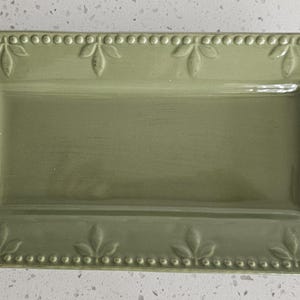 May include: A rectangular sage green ceramic serving tray featuring a raised beaded border and embossed leaf motifs along the edges. The tray has a smooth, glossy finish and a flat center, providing a functional surface for serving food or items.