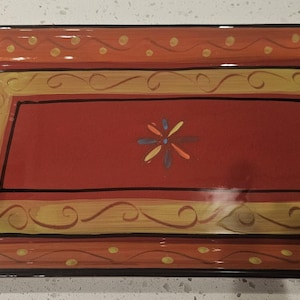 May include: A rectangular ceramic serving platter featuring a deep red center with a small multi-colored floral motif. The border is painted in shades of orange and yellow with a repeating pattern of dots and swirling lines in black.