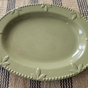 May include: An oval-shaped serving platter in a muted sage green color. The rim features a raised beaded texture interspersed with embossed leaf motifs at regular intervals. The surface has a smooth, glossy finish with a subtle sheen.