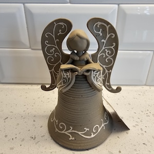 May include: A ceramic angel figurine in a seated position, reading a book. The angel is a muted gray color with white decorative scrollwork on the wings and base. The figurine has a bell-shaped body and is on a white surface.