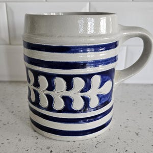 May include: A cylindrical ceramic mug featuring a light gray base color with deep blue horizontal stripes and a central embossed leaf pattern. The mug has a sturdy side handle and a smooth, glazed finish, showcasing a distinct contrast between the blue painted details and the neutral gray ceramic body.