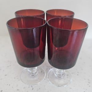 Op de afbeelding: Set van vier rode glazen met steel. De glazen zijn helder met een diepe rode tint en hebben een eenvoudig, elegant ontwerp.