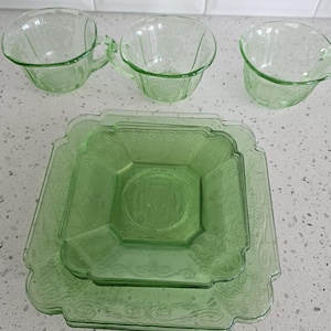 May include: A set of light green pressed glass tableware consisting of three cups with handles and two square plates. The glass features a decorative embossed pattern with floral motifs and geometric borders, showcasing a translucent finish.