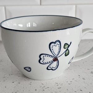 May include: A white ceramic mug featuring a hand-painted blue and green floral motif with a dark blue rim. The design includes a stylized three-petaled flower with dotted outlines and small green leaves, complemented by scattered blue petal accents.