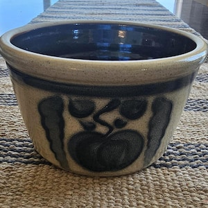 May include: A round, stoneware bowl with a wide rim and a dark blue interior. The exterior is a light beige with a dark blue fruit design. The bowl is likely used for serving or display.