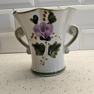 Può includere: Un vaso in ceramica color crema con bordo smerlato e due manici laterali. Il vaso presenta un disegno dipinto a mano di un fiore centrale viola con foglie verde scuro e piccoli punti marroni. Una fascia verde accentua la base e il bordo.
