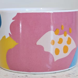Puede incluir: Un macetero de cerámica blanca con un patrón abstracto en rosa, amarillo y azul. El macetero tiene una forma redondeada y un acabado liso y brillante.