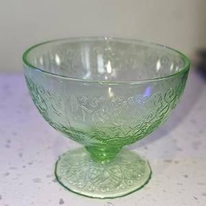 Può includere: Una coppetta da dessert in vetro verde della Depressione con base a piedistallo. La coppetta ha una forma arrotondata con disegni floreali e volute incise. Anche la base presenta dettagli incisi. Il vetro ha una leggera tinta verde.
