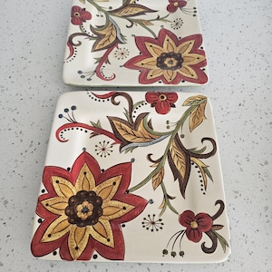 May include: Two square ceramic plates featuring a cream-colored base with a decorative floral pattern. The design includes large stylized flowers in shades of red, gold, and brown, accented with green leaves, swirling vines, and small dark blue dots.