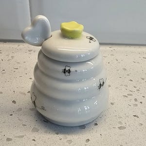 Puede incluir: Tarro de miel de cerámica blanca con forma de colmena, con asa en forma de corazón y pomo en forma de flor amarilla. El tarro está decorado con ilustraciones de abejas negras.