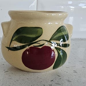 Puede incluir: Una vasija de cerámica color crema con una abertura ancha y dos pequeñas asas laterales. El exterior presenta un diseño pintado a mano de una manzana de color rojo oscuro con hojas y tallos verdes que rodean el cuerpo redondeado.