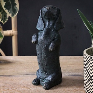 May include: A black dachshund figurine standing on its hind legs with its front paws raised. The figurine is made of a dark, textured material and has a realistic appearance.