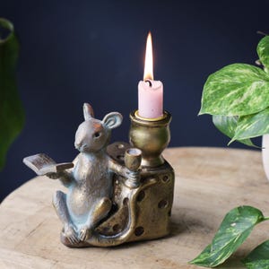 May include: A bronze-toned candle holder featuring a sculpted mouse holding an open book and a small goblet. The mouse sits beside a cylindrical candle cup containing a lit pink candle. The base is shaped like a block of Swiss cheese with holes.