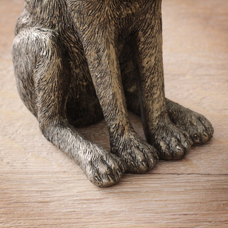 May include: A detailed view of a dog statue's front paws and lower legs. The statue has a textured, metallic finish in grey and gold tones. The dog is positioned on a wooden surface.