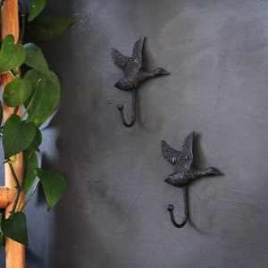 May include: Two black metal hooks shaped like flying ducks. The hooks are mounted on a gray wall.
