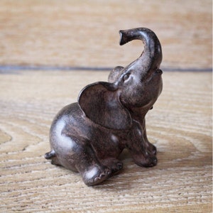 May include: A small, brown elephant figurine with its trunk raised. The elephant is sitting on its hind legs, with large ears and detailed feet. The figurine is made of a dark, textured material and sits on a light brown wooden surface.