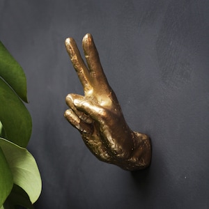 Könnte beinhalten: Eine goldfarbene Metallskulptur einer Hand, die das Friedenszeichen macht. Die Hand ist erhoben und leicht nach rechts geneigt.