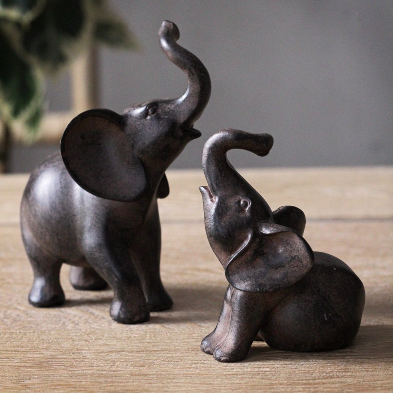 May include: Two dark brown elephant figurines. One stands with its trunk raised, and the other is seated with its trunk up. The elephants have large ears and are placed on a light brown wooden surface. The background is a neutral gray.