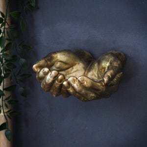 May include: A golden-colored wall sculpture depicting two cupped hands. The hands are detailed with visible knuckles and lines, suggesting a textured surface. The sculpture is mounted on a dark blue wall, with green foliage visible on the left side.