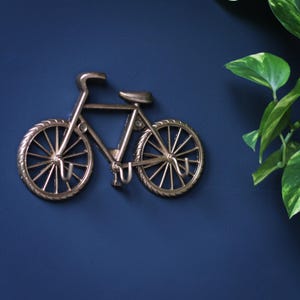 Pode incluir: Um gancho de parede em metal cor de bronze em forma de bicicleta. O gancho está montado numa parede azul escuro, com uma planta verde no fundo. A bicicleta tem dois ganchos para pendurar objetos.