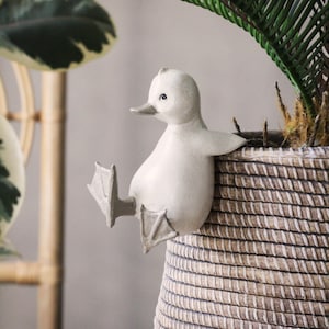 May include: A white ceramic duck figurine perched on the edge of a woven wicker planter. The duck has a long beak and webbed feet.