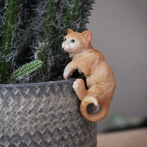 May include: A small orange figurine of a cat climbing up the side of a grey planter. The cat has blue eyes and a white patch on its chest.