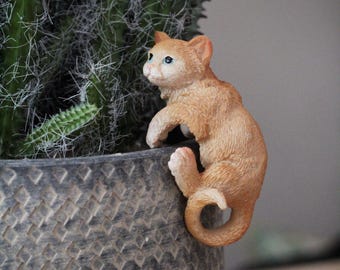 Puede incluir: Una pequeña figurita naranja de un gato subiendo por el lado de una maceta gris. El gato tiene ojos azules y una mancha blanca en el pecho.